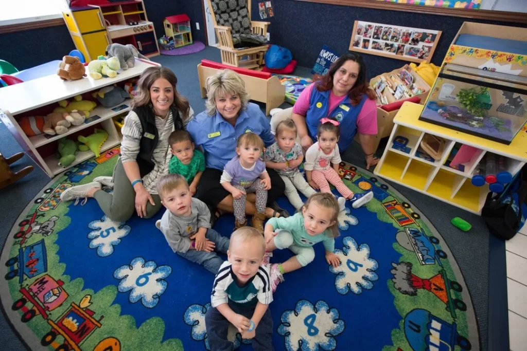 Three credentialed preschool teachers sitting with a group of young children on a colorful classroom rug surrounded by organized interest areas at ABC Child Development Center West in Greeley.