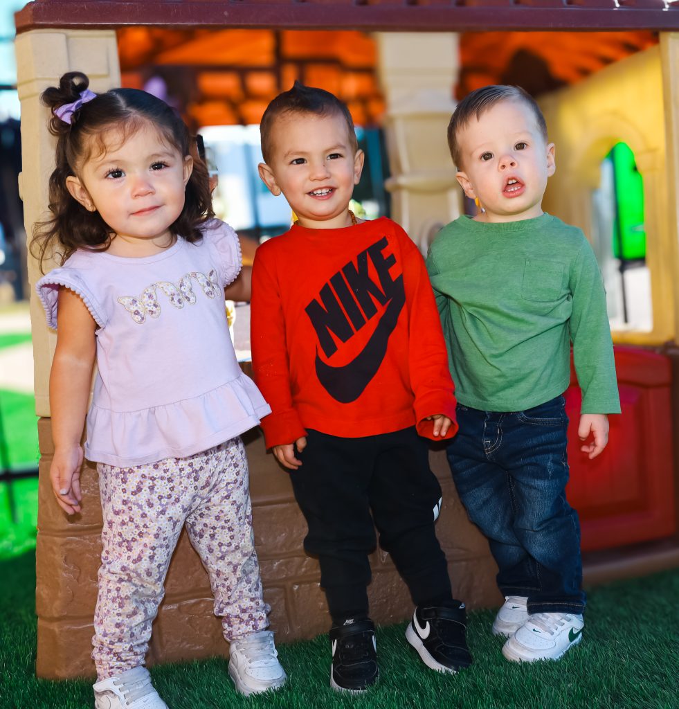 Three toddlers standing together and smiling outside a playhouse at ABC Child Development Center in Ault, Colorado